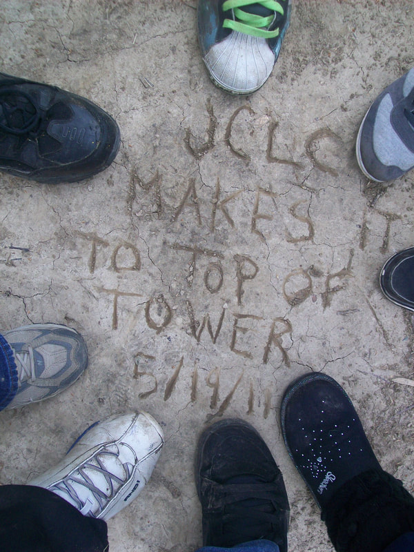 Overhead view of several student's shoes. In the middle of the circle of feet is a message written in the dirt that says "We made it to the top of the Elba Tower"