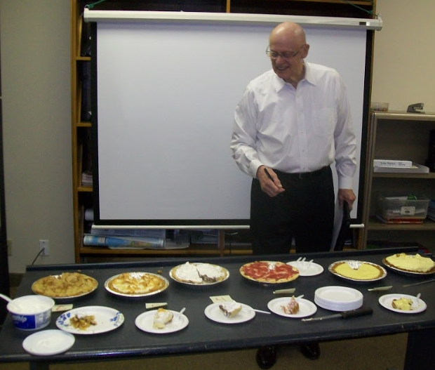 A photo of a man standing in front of 12 student made pies for a pie making contest. The contest was a fundraiser for school travel.