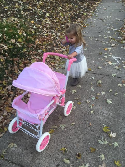 A little girl walking her baby doll's stroller down the street with a toy cell phone in her hand.