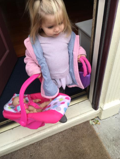 A little girl walking with her baby doll in a carrier, with her toy purse in her other hand.