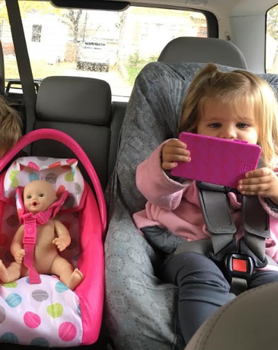 A little girl sitting in her car seat with a toy cell phone in her hand and her baby doll strapped into the car seat next to her.